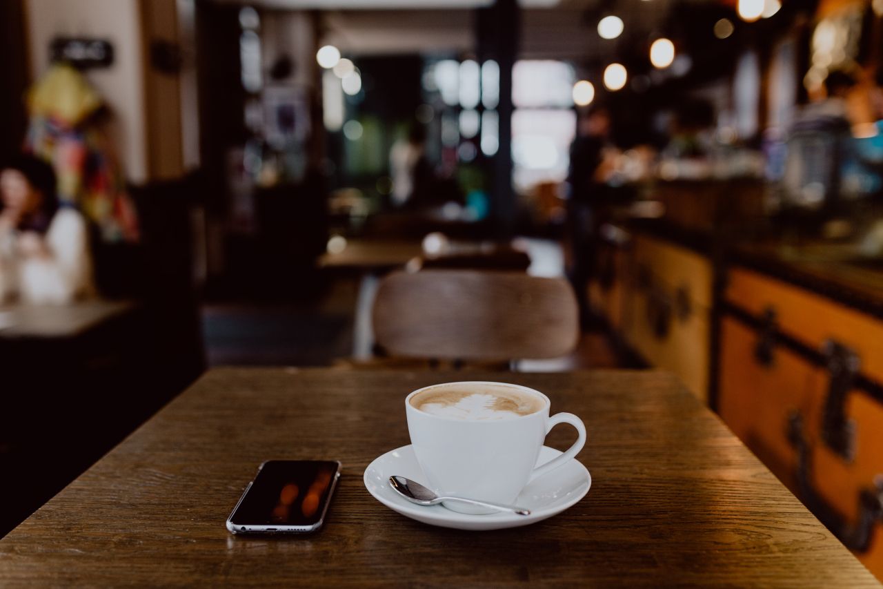 A cup of coffee on a wooden cafe table with a smartphone lying beside it.